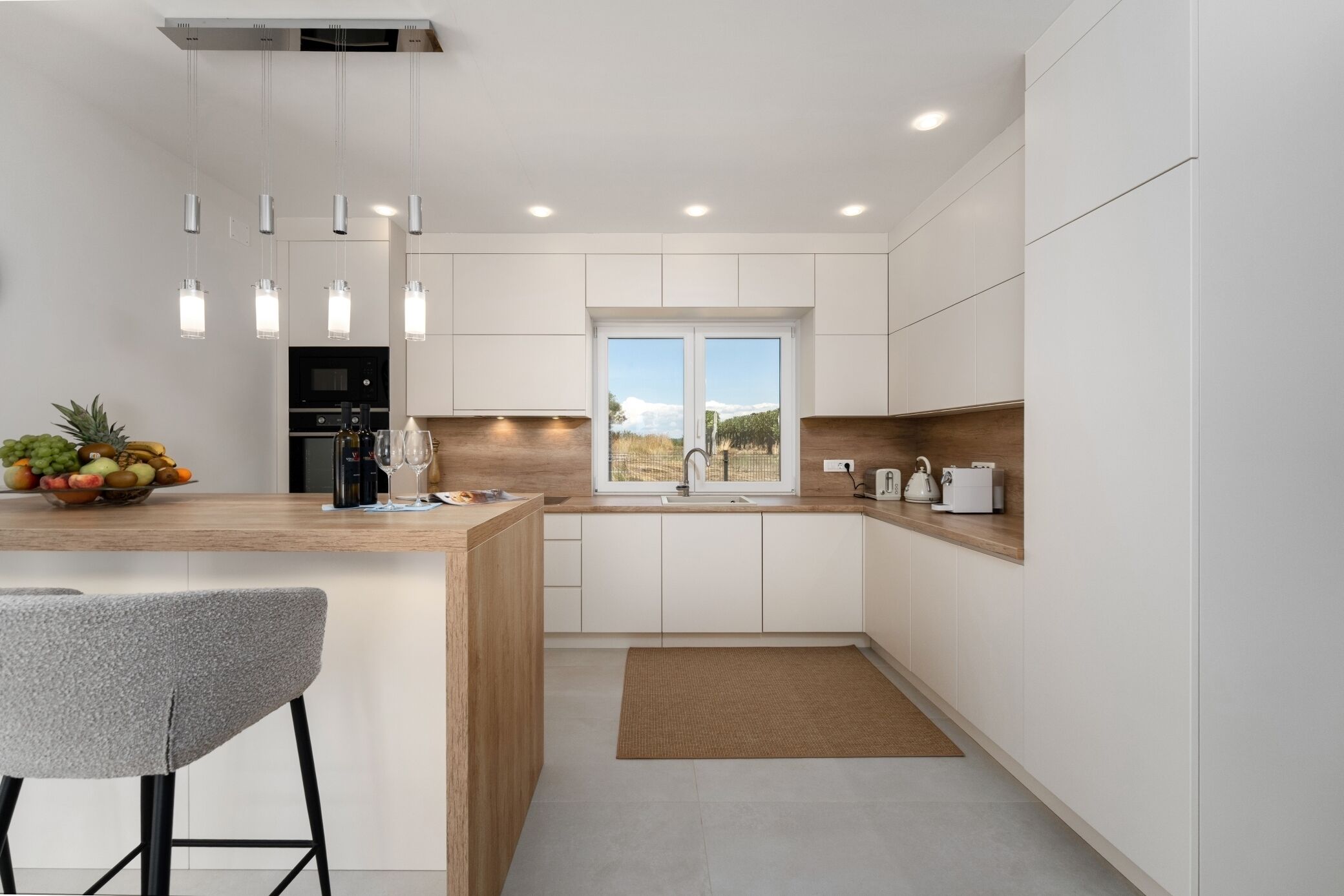A modern kitchen with white cabinets, a wooden countertop, a built-in island with a bar stool, a window with a view, and recessed lighting.