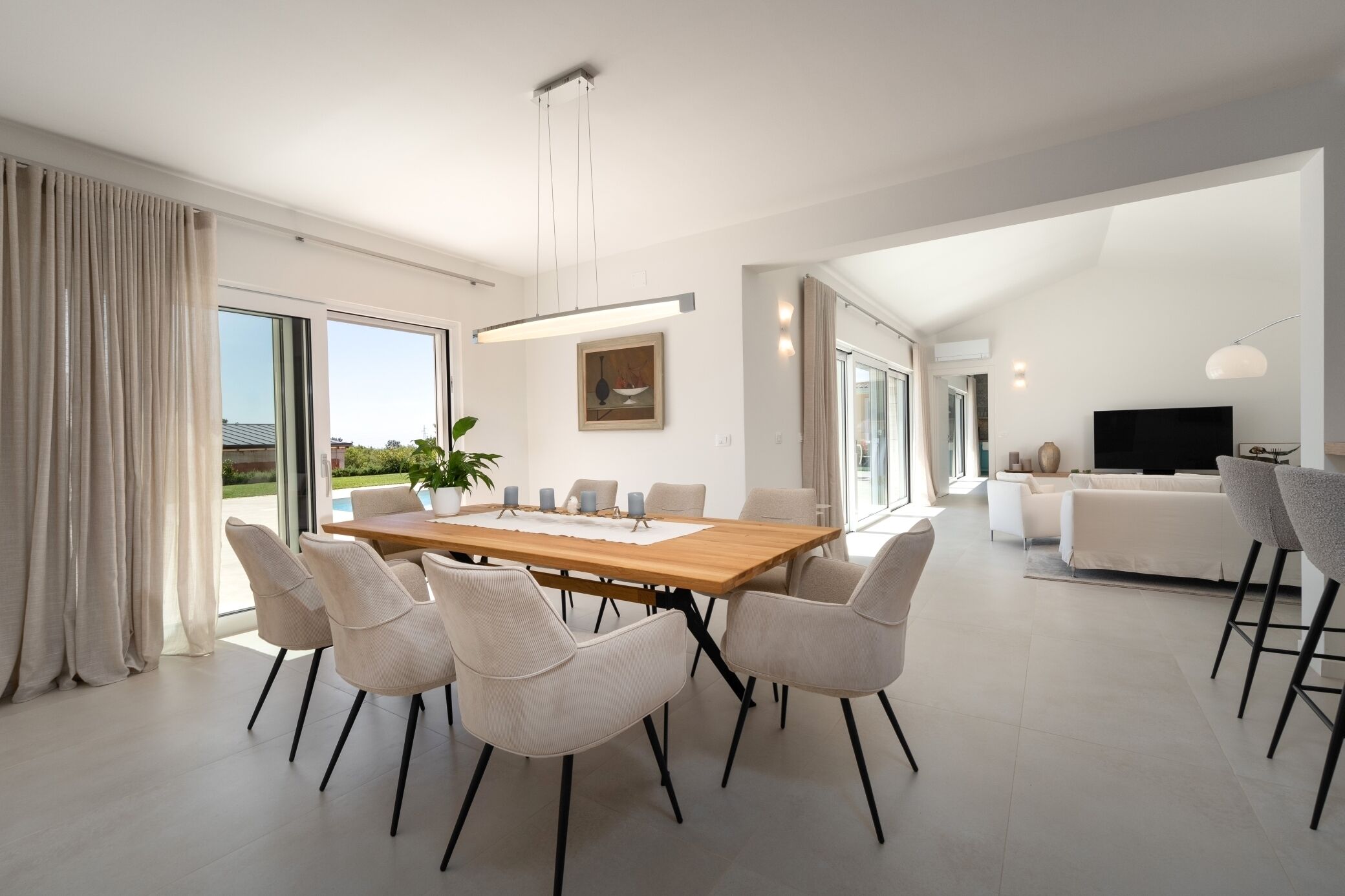 Een moderne eetruimte met een houten tafel en stoelen, een groot raam met gordijnen, en een flatscreen televisie op de achtergrond.