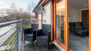 En balkon med to fletstole, en skydedør der fører til en stue med sofa og mønstrede hynder, samt en parabolantenne på væggen.