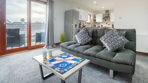 En moderne stue med en grå sofa, et Monopoly brætspil og udsigt til bybilledet gennem store vinduer.