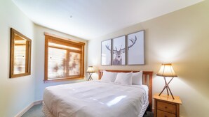 A neatly arranged bedroom with a large bed, two bedside tables with lamps, a window with blinds, and three framed pictures on the wall. - Ski in/Out 1B/1BA w/ View - Pool, Jacuzzi, Parking (Mammoth Lakes)