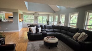 A spacious living room with a large sectional sofa, a round ottoman, and a wooden floor. There is a skylight above the sofa and a kitchen area visible through a glass door. - Beautiful mountain views for a fun, relaxing getaway! (Seven Devils)
