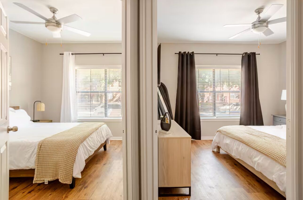 Une vue en miroir d’une chambre d’hôtel avec deux lits, un plancher en bois, des ventilateurs de plafond et une fenêtre avec des stores.