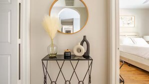 A bedroom with a bed, a bedside table with a mirror, a vase with dried flowers, and a small candle.