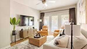 A living room with a flat-screen TV, a ceiling fan, a sofa, a coffee table, and a potted plant.