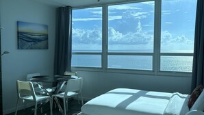 Una habitación de hotel con un ventanal que ofrece vistas al mar y nubes, una cama, un escritorio con una silla y un cuadro en la pared.