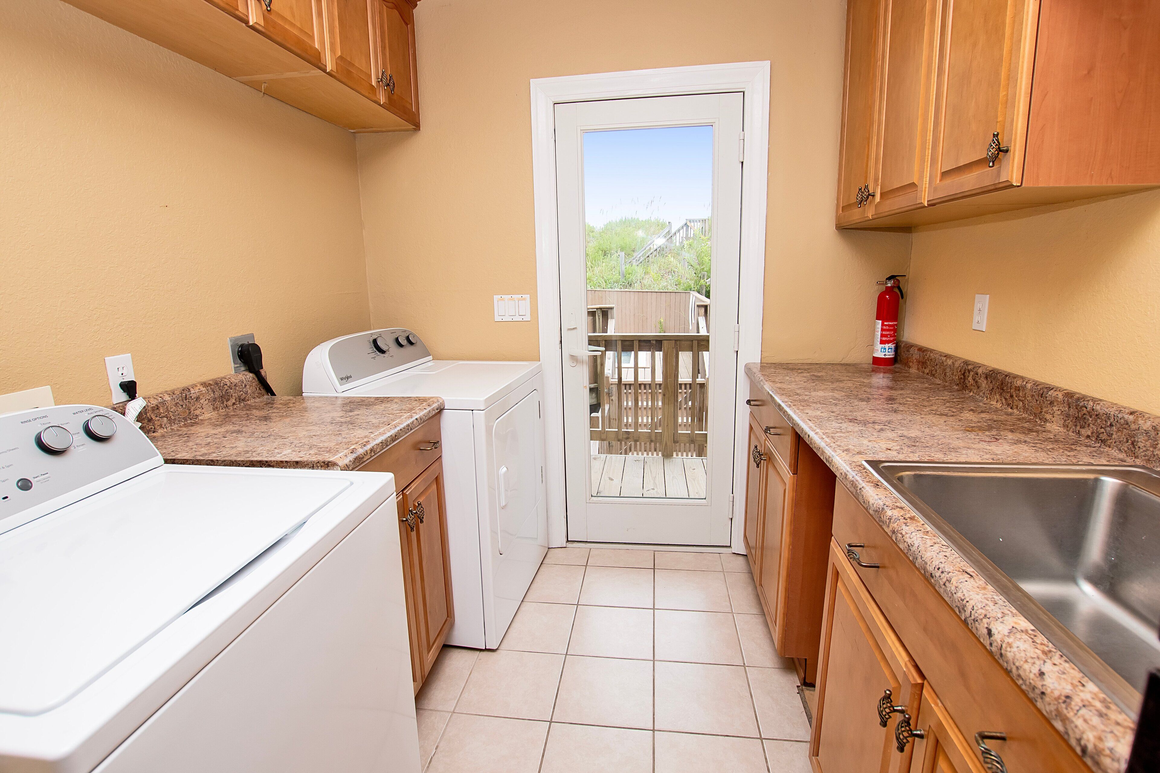 A compact kitchen with a washing machine, a dryer, a stainless steel sink, wooden cabinets, and a door leading to a balcony.