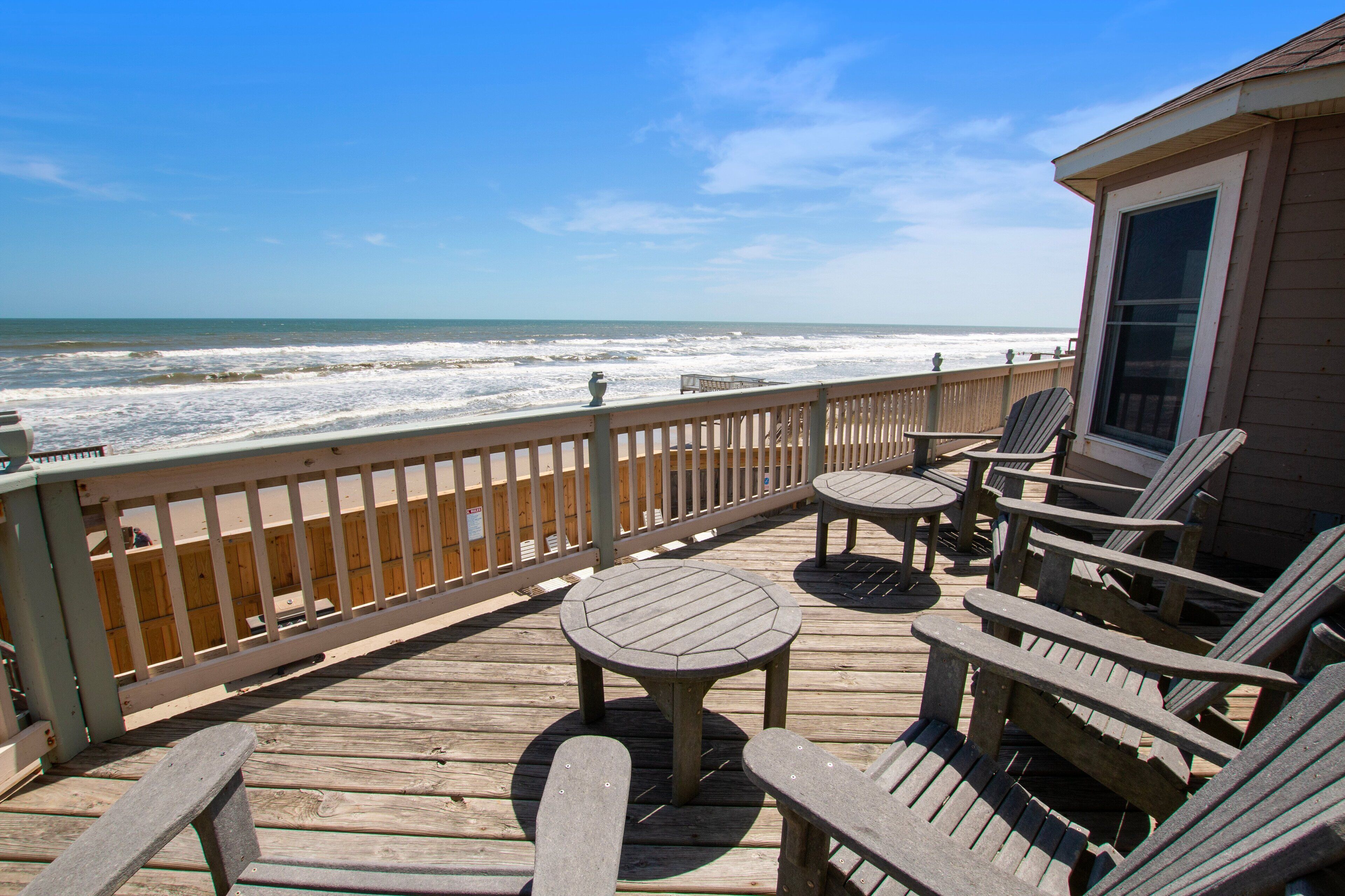 Um deck de madeira com cadeiras Adirondack e uma mesa, com vista para uma praia com ondas.