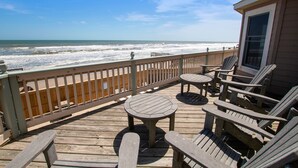 Une terrasse en bois avec des fauteuils Adirondack et une table, donnant sur une plage où l’on peut admirer les vagues.