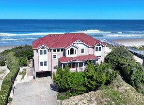 A two-story house with a red roof, located near a beach with a wooden walkway.