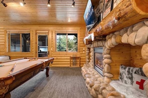 A log cabin interior with a pool table, stone fireplace, and large windows.