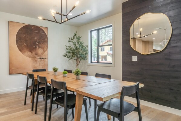 A modern dining area with a wooden table, black chairs, a large abstract painting, a potted plant, and a round mirror on the wall.