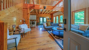 Una cabaña de madera espaciosa con un dormitorio entrepiso, una sala de estar con sofá y sillones, y una cocina al fondo.