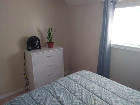 A bedroom with a bed, a dresser, a potted plant, a fan, and a window with curtains.