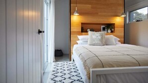 Un dormitorio moderno con cabecera de madera, una cama grande con ropa blanca, lámparas colgantes y vistas al exterior a través de una ventana.