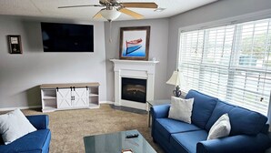 Una sala de estar con un sofá azul, una mesa de centro de vidrio, una chimenea, un ventilador de techo y un televisor montado en la pared.