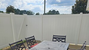Una mesa de patio con seis sillas, una cerca blanca y árboles al fondo.