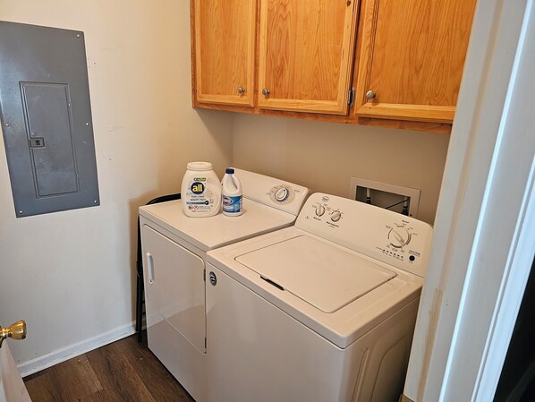 A laundry room with a front-loading washer and dryer, wooden cabinets above, and a gray door on the left.