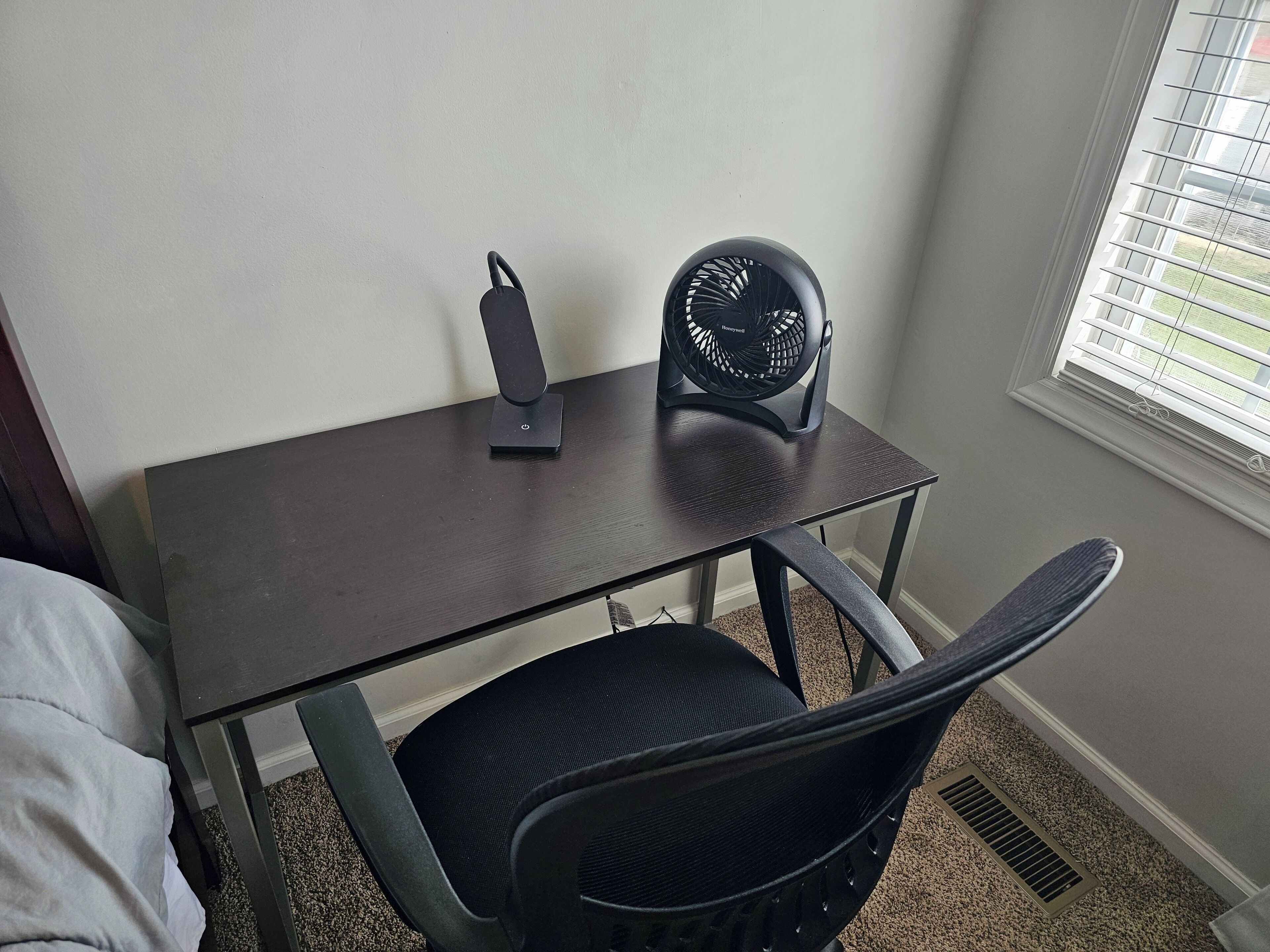 Un petit bureau en bois foncé, avec un ventilateur noir et une chaise noire.