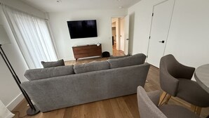 Un salon moderne avec un canapé gris, un téléviseur fixé au mur, un plancher en bois et une porte donnant sur une autre pièce.
