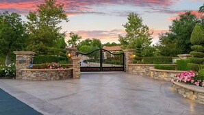 A gated entrance with stone walls and a metal gate, surrounded by well-maintained hedges and flowering plants.