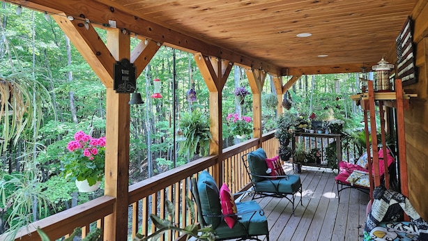 A wooden deck with two green chairs and a table, surrounded by lush greenery and hanging plants.