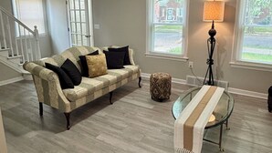 Una sala de estar con un sofá color beige, una mesa de centro de vidrio y una lámpara. Hay ventanas con persianas y una escalera al fondo.