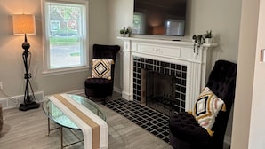 Una sala de estar con chimenea, dos sillones, una mesa de centro de vidrio y un televisor montado sobre la chimenea.