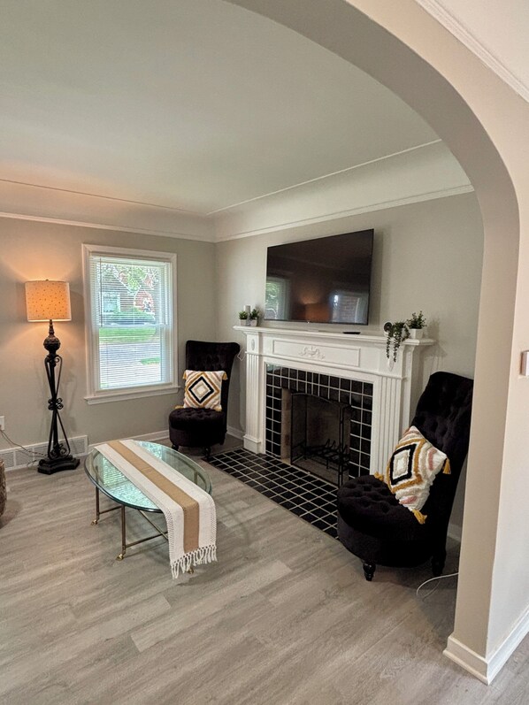A living room with a fireplace, two armchairs, a glass coffee table, and a TV mounted above the fireplace.