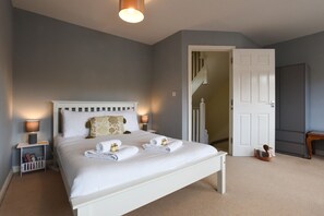 Een net opgemaakt bed met witte lakens en handdoeken, een nachtkastje met een lamp, en een deur naar een trap.
