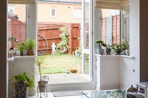 Uitzicht vanuit een kamer met een groot raam op een achtertuin. Op de vensterbank staan potplanten en in de tuin is een houten omheining en een beeld te zien.