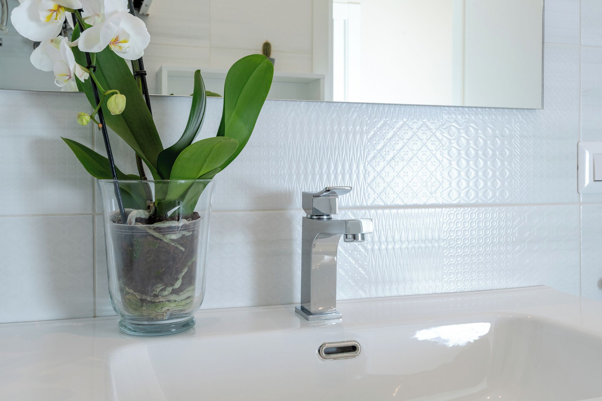 Een badkamer met een witte wastafel, een moderne kraan en een glazen vaas met witte orchideeën.