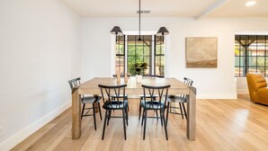 Un espace repas moderne avec une table en bois et des chaises noires, de grandes fenêtres donnant sur un paysage verdoyant, et un fauteuil jaune dans un coin.