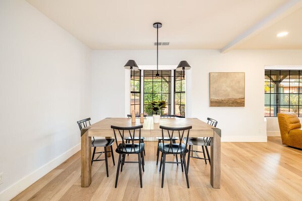 A modern dining area with a wooden table and black chairs, large windows with a view of greenery, and a yellow armchair in the corner.
