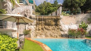 A swimming pool with a waterfall feature, surrounded by a stone wall and lush greenery.