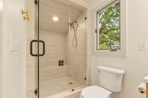 Un baño moderno con ducha separada por mampara de vidrio, inodoro y ventana con vistas a árboles.