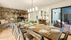 A dining area with a long wooden table set for six, a stone fireplace, and a view of the outdoors. - Family Base Camp + 5BR + Game Room + Hot Tub (McGaheysville)