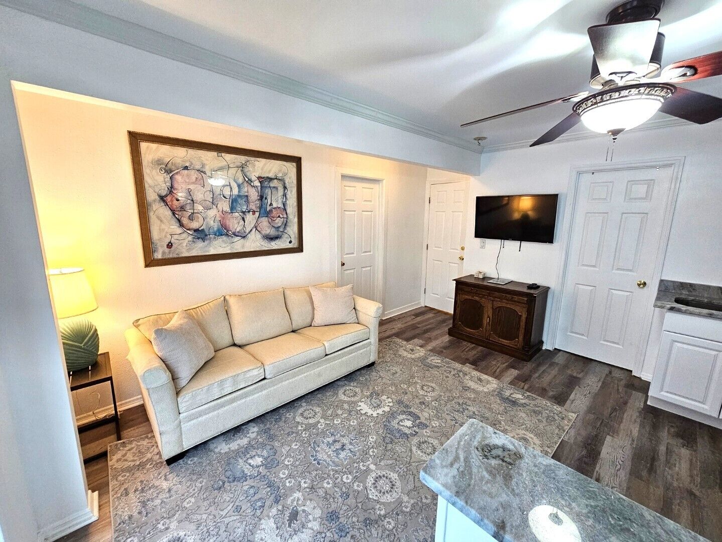 A living room with a beige sofa, a large abstract painting, a ceiling fan, and a flat-screen TV.