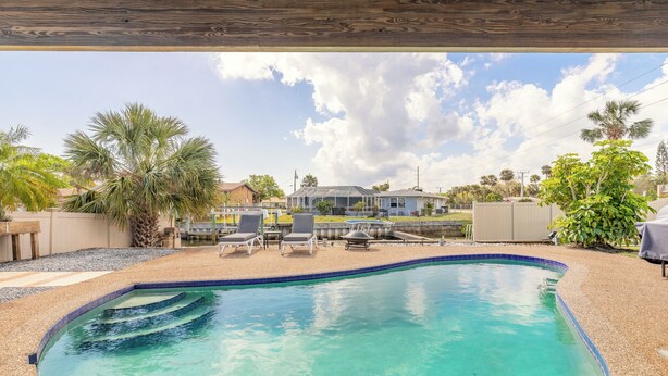 A swimming pool with a wooden deck overhead, surrounded by palm trees and a house in the background.