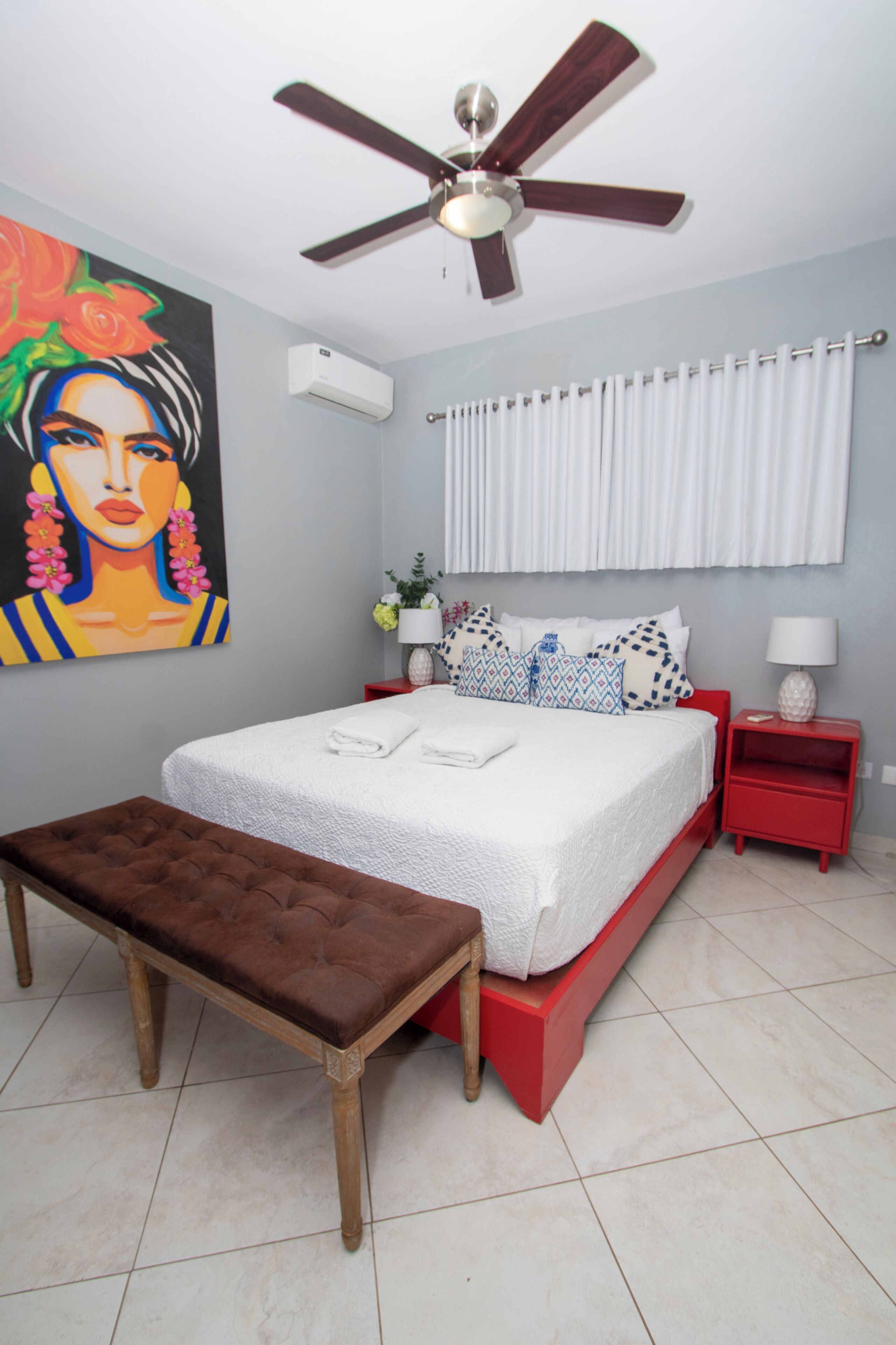 A bedroom with a bed, a red nightstand, a white air conditioner, a ceiling fan, a bench, and a painting on the wall.