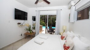 A hotel room with a bed, a TV, a ceiling fan, a window with curtains, and a view of an outdoor pool. - Luxury Caribbean 5BR Villa – Private Pool, BBQ & Ocean Breezes (Sosúa)