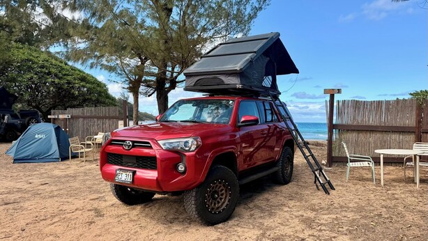Un SUV rojo con una tienda de campaña en la azotea sobre una playa arenosa.
