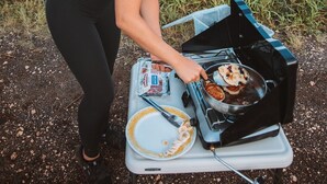 Una persona cocina al aire libre usando un fogón portátil, rodeada de paisajes naturales.
