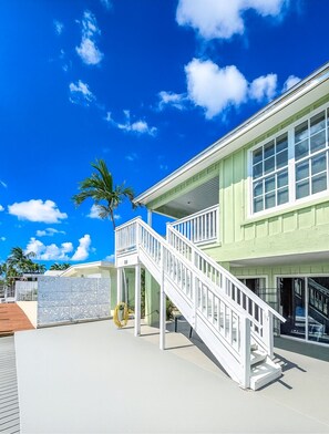 A two-story building with a white staircase leading to a balcony, a palm tree, and a clear blue sky. - Welcome to Castillo Del Mar. A peaceful, salty newly remodeled ocean front home! (Key Colony Beach)