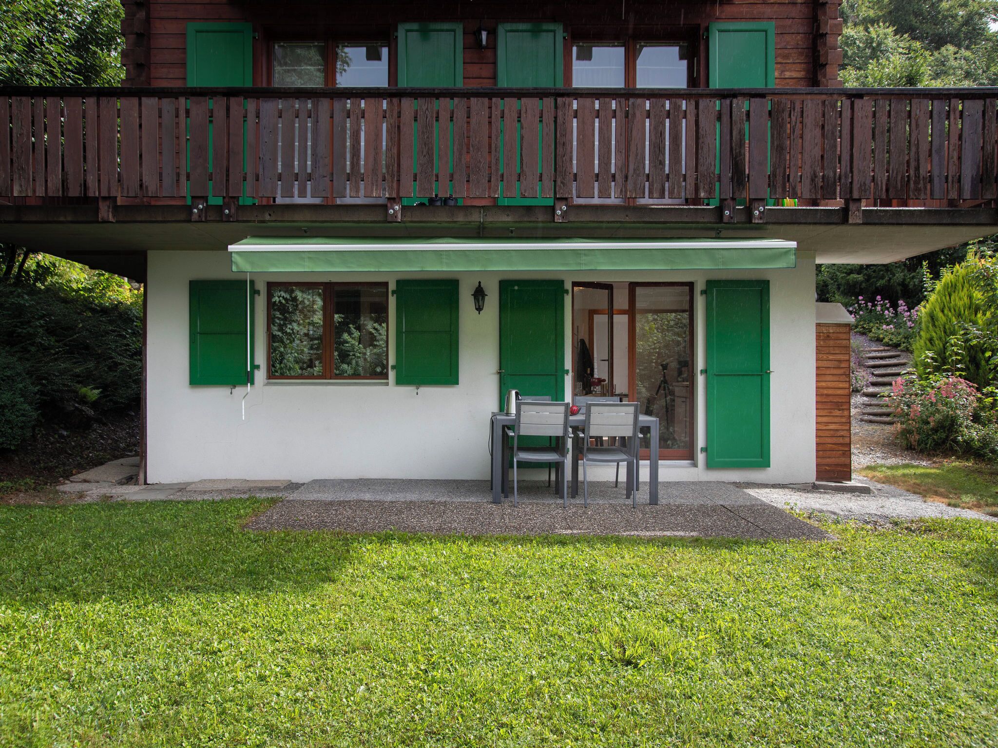 Una casa de fusta de dos pisos amb persianes verdes, un balcó i una petita zona d'estar a l'aire lliure amb taula i cadires.