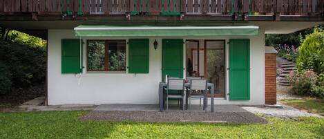 A two-story wooden house with green shutters, a balcony, and a small outdoor seating area with a table and chairs.