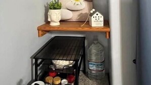 A corner with two wooden shelves, one holding a potted plant, a decorative item, and a framed picture with a daisy. Below, a black metal shelf contains various items and a water bottle.