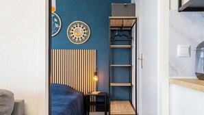 Quarto compacto com cama, mesa de cabeceira iluminada por um abajur, guarda-roupa e um pequeno espaço de estar.