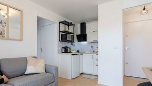 Um apartamento moderno com um sofá cinzento, uma pequena mesa redonda, uma cozinha com armários brancos e uma estante preta para televisão.