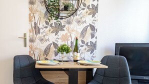 Uma área de refeições com uma mesa posta para dois, com papel de parede floral, duas cadeiras cinzas estofadas, uma pequena planta em vaso, uma garrafa de vinho e uma televisão sobre um suporte de madeira.